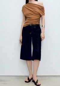 Brun off-the-shoulder top med samlet tekstur; parret med mørkeblå knælange denim culotter og brune flip-flops.