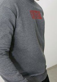Sudadera gris con mangas largas, cuello redondo y texto rojo "COUTURE" impreso en la parte delantera. La tela parece suave con una textura lisa.