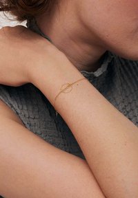 Złota bransoletka łańcuszkowa z okrągłym akcentem i małym charms, noszona na jasnej karnacji na tle teksturowanej szarej tkaniny.