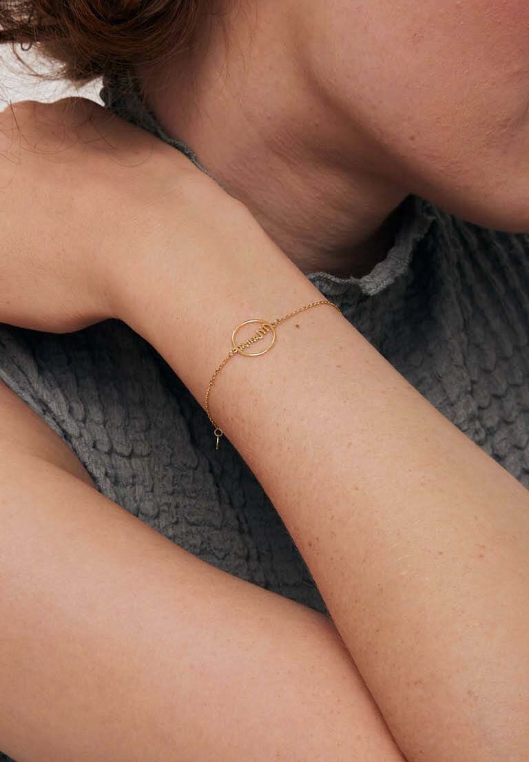 Złota bransoletka łańcuszkowa z okrągłym akcentem i małym charms, noszona na jasnej karnacji na tle teksturowanej szarej tkaniny.