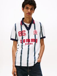 Weißes Poloshirt mit vertikalen blau-grünen Streifen, roter "85" und "Tommy Jeans" Logo, marineblauer Kragen und kurzen Ärmeln. Glatte Textur.