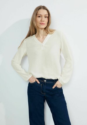 Blusa bianca a maniche lunghe con scollatura a V e tessuto texturizzato, abbinata a jeans in denim blu scuro. La blusa presenta un piccolo dettaglio con bottone.