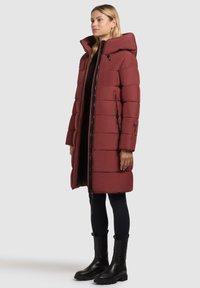 Abrigo de plumas en rojo intenso con un cuello alto, cremallera completa y bolsillos laterales. Presenta un diseño acolchado y se combina con leggings negros y botas.