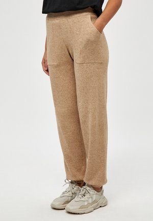 Personne portant un pantalon large en tricot beige avec des poches devant et des baskets gris clair sur un fond blanc uni.