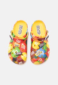 Sabots colorés sur le thème des M&M's en matériau plastique, présentant un design multicolore, des logos en relief et des trous de ventilation pour la respirabilité.