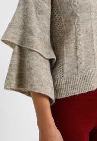 Pull gris tricoté avec des manches larges évasées, présentant un motif torsadé et un ourlet côtelé. Texture douce et tissu chaud, associé à un pantalon rouge foncé.