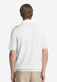 Vit piké med korta ärmar, texturerat tyg och ett ribbat krage. Anmärkningsvärda axelaccenter i en kontrasterande färg.