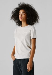 Žena s kudrnatými vlasy, oblečená v světle šedém tričku s malým nápisem na hrudi a černých kalhotách, stojící s jednou rukou v kapse.