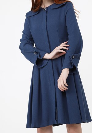 Femme portant un manteau bleu marine mi-long à manches longues avec des détails décoratifs à boucle sur les poignets et le col.