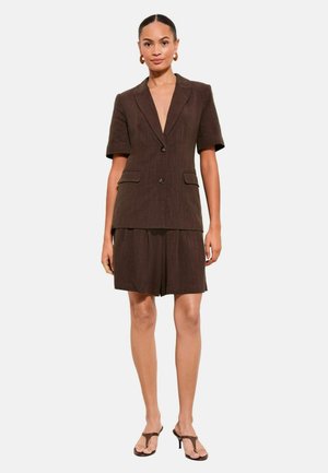 Vrouw draagt een bruin kort mouwblazer met knopen en bijpassende knielange shorts, gecombineerd met bruine sandalen met hak en oorringen in de vorm van hoepels.