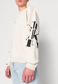 Personne portant un pull crème avec des lettres abstraites noires sur la manche et un jean bleu clair sur fond uni.