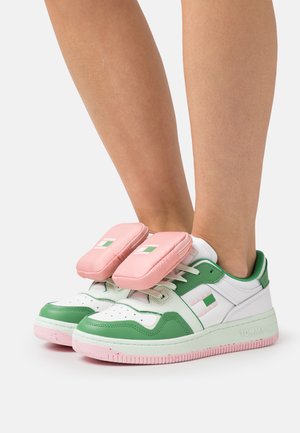 Witte en groene sneakers met roze zolen gedragen, voorzien van verwijderbare roze kussentjes die over de veters zijn bevestigd.