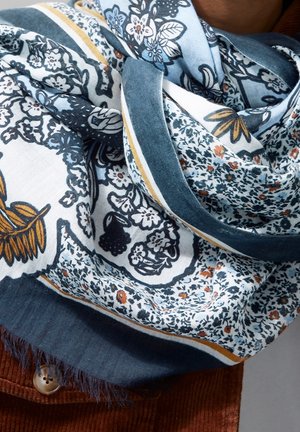 Écharpe en coton doux avec des motifs floraux et de feuilles bleus, blancs et moutarde, enroulée autour d'une personne portant une veste en velours côtelé marron avec des boutons.