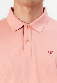Hellrosa Polohemd mit strukturierter Oberfläche, ausgestattet mit einem Kragen, zwei Knöpfen und einem kleinen bordeauxroten Logo auf der Brust.