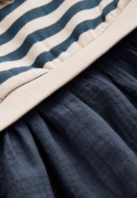 Stribet stof i creme og mørk teal, med en ribbet beige kant, lagdelt over blødt, tekstureret mørkeblåt stof.