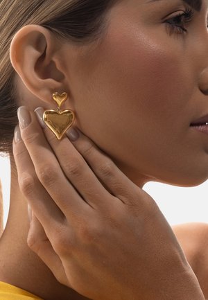 Zbliżenie na kobietę dotykającą szyi, noszącą złote, podwójne kolczyki w kształcie serca, z gładką skórą i neutralnym lakierem do paznokci.