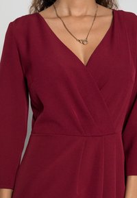 Robe bordeaux avec un décolleté en V et des manches longues, présentant un design drapé et une texture lisse. Un collier en chaîne dorée fine met en valeur le cou.
