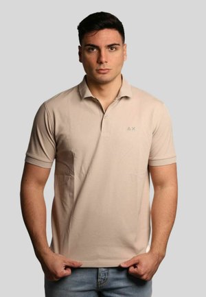 Polo beige con collo, maniche corte e un logo sottile in verde scuro. Stile con orlo dritto e superficie liscia.