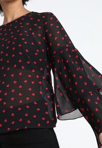 Blouse noire transparente avec motif de cœurs rouges, manches larges en cloche et col rond. Tissu léger offrant une texture douce.
