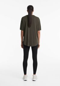 Oliven grøn oversized t-shirt med korte ærmer, buet kant, båret med sorte leggings og hvide sportssko. Glat tekstur, afslappet design.