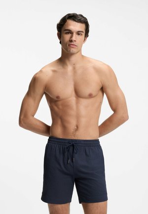 Young man with dark hair standing shirtless, wearing dark blue drawstring swim shorts, hands behind his back, neutral expression.