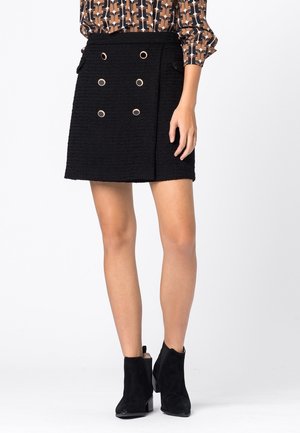 Kobieta ubrana w czarną teksturowaną mini spódniczkę z sześcioma ozdobnymi złotymi guzikami, w połączeniu z czarnymi botkami i wzorzystą bluzką.