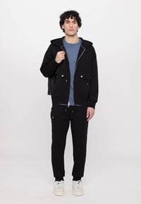 Sudadera con cremallera negra y capucha con cordón, bolsillos frontales; combinada con una camiseta azul y pantalones de chándal cargo negros, junto con zapatillas blancas.