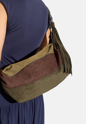 Kobieta ubrana w granatową sukienkę, nosząca oliwkowo-brązową zamszową torbę na ramię z dużym frędzlem na pasku.