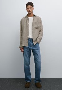 Camicia beige a scacchi, t-shirt bianca a girocollo, jeans dritti azzurri chiari, scarpe in suede marroni, in piedi contro uno sfondo semplice.