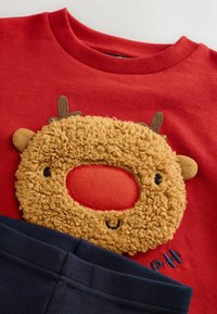 Czerwony sweter z puszystą, teksturowaną łatą w kształcie zwierzęcej twarzy, z brązowymi rogami i wyszytym uśmiechem, w połączeniu z granatowymi spodniami.