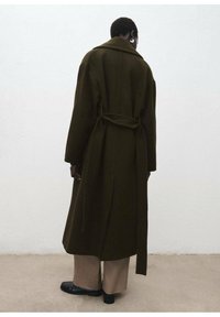 Manteau long en laine vert olive avec une taille ceinturée, un large col et des épaules basses, porté sur un pantalon large beige et des chaussures noires.