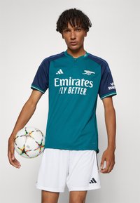 Maglietta da calcio color turchese con accenti blu navy, presenta il logo bianco "Emirates" e il marchio "adidas". Il modello tiene in mano un pallone da calcio colorato. Pantaloni bianchi.