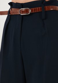 Pantaloni bleumarin cu o croială ajustată, detaliu cu plisat și o curea din piele de crocodil maro cu o cataramă ovală argintie. Textură de material neted.
