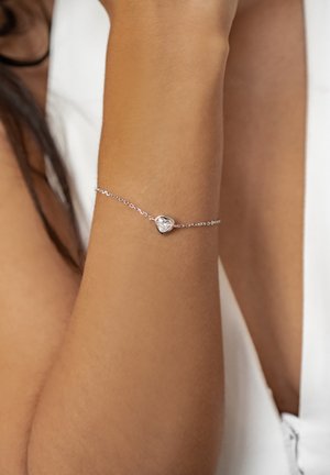 Delicate silver chain bracelet with a small heart-shaped charm featuring a clear gemstone, worn on a tanned wrist against white clothing.