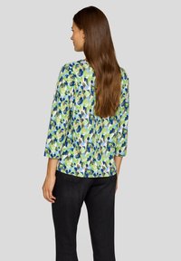 Bluse mit floralem Muster in Grün-, Blau- und Weißtönen. Mit Dreiviertelärmeln und lockerer Passform. Rückansicht; getragen mit dunklen Hosen.