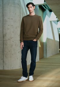 Ruskea college-paita, jossa on pyöreä kaula-aukkko, tummansiniset farkut ja valkoiset tennarit, joissa on kumipohjat. Malli seisoo modernissa sisätilassa.