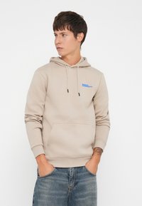 Beige hoodie with adjustable drawstrings, front kangaroo pocket, and blue logo text. Soft fabric with ribbed cuffs. Worn with faded jeans.