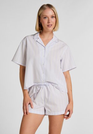 Vrouw met blond bobkapsel draagt een wit kort mouw shirt met knopen en bijpassende short met strikkoord en subtiele verticale strepen, staand.