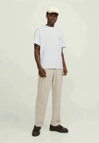 Jonge zwarte man die staat, gekleed in een witte pet, lichtblauw T-shirt, beige broek en donkerbruine schoenen tegen een effen lichte achtergrond.