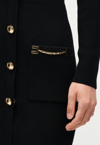 Μαύρο πλεκτό ζακέτα με χρυσά κουμπιά και διακοσμητική χρυσή αλυσίδα που συνδέει δύο τετράγωνα κλιπ σε μια μπροστινή τσέπη, δείχνοντας κοντινή εικόνα του χεριού.