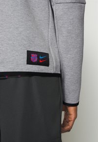 Sudadera gris con parche negro que presenta los logotipos de Barcelona y Nike. Tejido suave con puños acanalados. Usada sobre pantalones cortos oscuros.