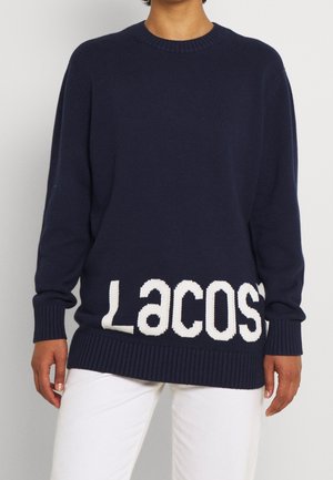 Personne portant un pull bleu marine avec un grand texte blanc "Lacoste" près de l'ourlet, associé à un pantalon blanc sur un fond uni.