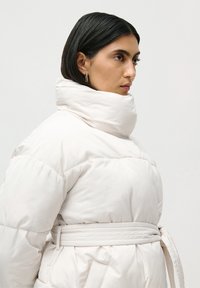 Femeie cu păr drept și negru, purtând o geacă puf albă cu centură și guler înalt, îndreptată spre dreapta, pe un fundal deschis.