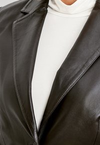 Czarna skórzana marynarka z klapami i wyraźnym kołnierzem, o gładkiej fakturze i przemyślanej sylwetce. Noszona na białym topie z golfem.