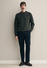 Dunkelgrüner Grobstrickpullover mit Rundhalsausschnitt, kombiniert mit navyblauen Hosen und dunklen Slippern mit goldenen Akzenten. Glatte Textur und taillierter Schnitt.