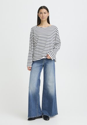 Mujer de pie vistiendo una camiseta de manga larga a rayas blancas y negras y pantalones vaqueros anchos azules con zapatos negros sobre un fondo liso.