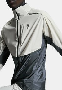 Veste de course légère en crème et gris foncé. Dotée d'un col haut, d'une fermeture éclair et de sections ventilées avec un motif texturé. Logo visible.