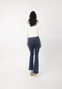 Jeans de mezclilla azul de corte acampanado con un lavado oscuro, presentan una cintura alta, dos bolsillos traseros y una textura sutil. Llevados con una blusa blanca de manga larga.