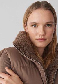 Braune gepolsterte Weste mit einem gemütlichen, strukturierten Kragen. Verfügt über einen Reißverschluss und ein glattes äußeres Material. Das Modell zeigt das Kleidungsstück vor einem neutralen Hintergrund.