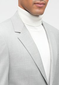 Blazer cinza claro com um padrão xadrez subtil, combinado com um suéter de gola alta branco. O tecido parece liso e bem ajustado.